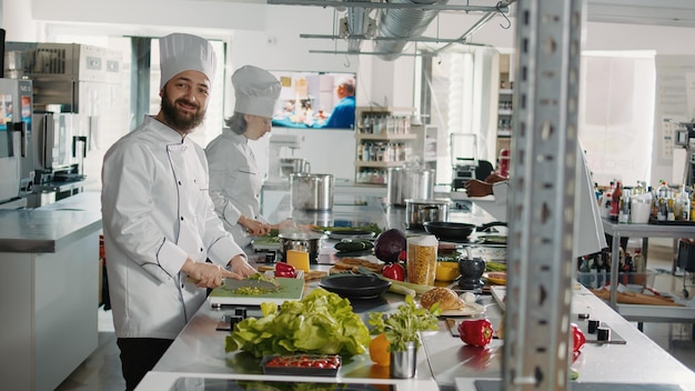 Portrait of man in uniform preparing green salad on cutting board to cook gastronomy cuisine dish for restaurant menu. Professional chef working on culinary recipe with organic food. Handheld shot.