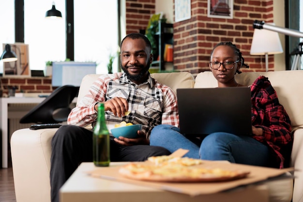 Portrait of man watching tv film and woman working on laptop, browsing internet and having fun with movie on television. Eating fast food meal from takeout delivery and drinking bottles of beer.