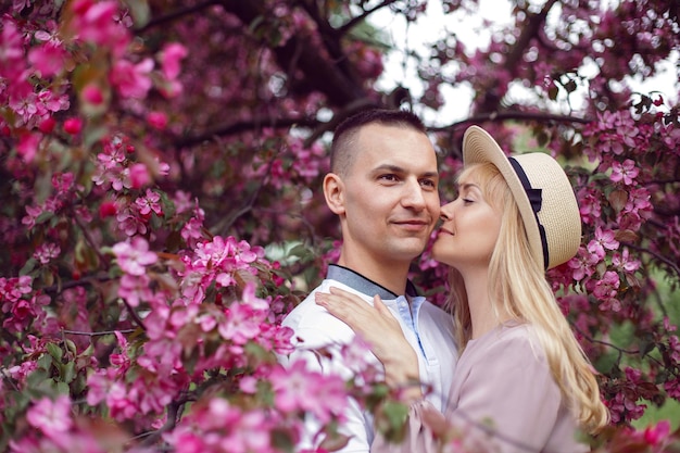 Portrait man and a woman in hat stand by a blooming pink cherry tree in summer