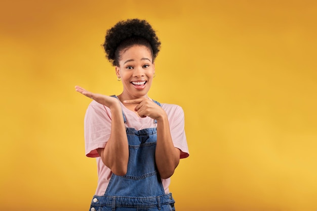 Portrait marketing and a black woman pointing to her palm for the promotion of a product on a yellow background in studio Smile advertising or space with a happy young female brand ambassador