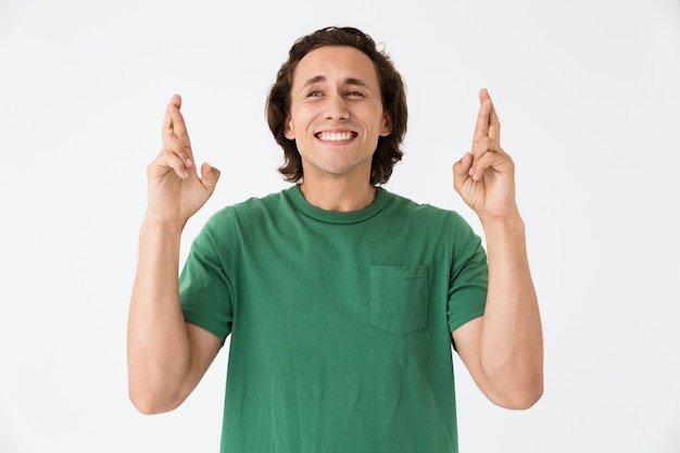 Portrait of masculine young man wearing basic t-shirt keeping fingers crossed isolated over white wall