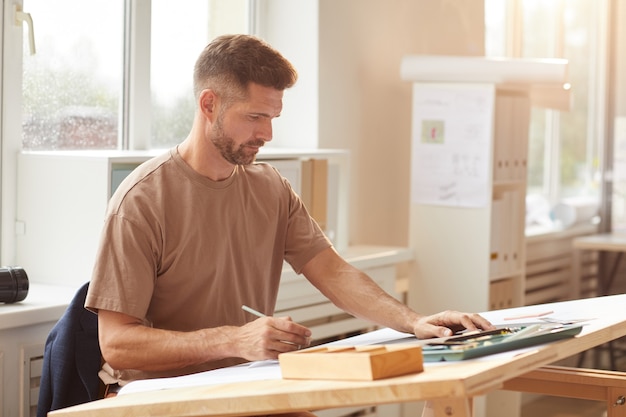 Portrait of mature bearded architect looking at blueprints while sitting at drawing desk in sunlight, 