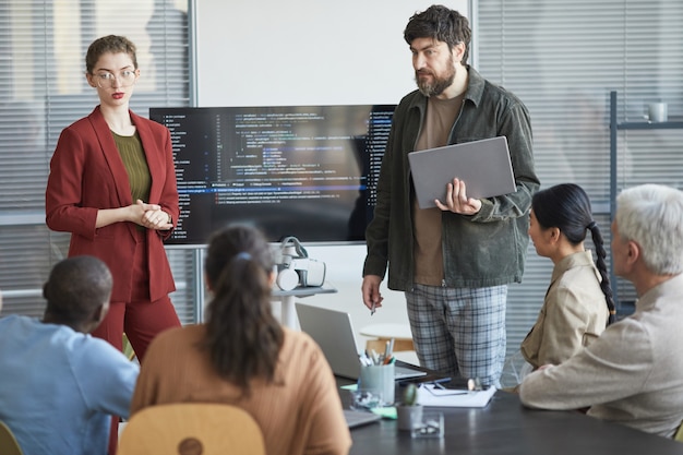 Portrait of mature bearded man giving presentation in office to diverse IT team standing by code on screen