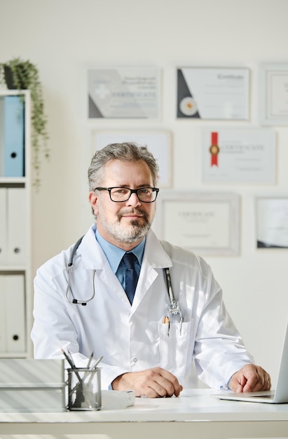 Portrait of mature doctor in white coat sitting at his workplace at office