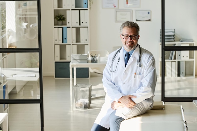 Portrait of mature doctor in white lab coat smiling at camera while working at office