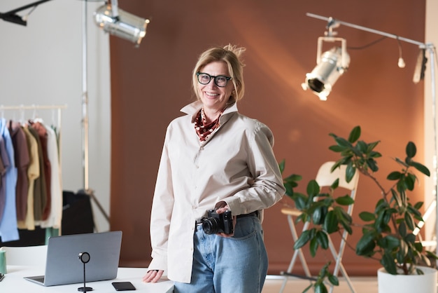 Portrait of mature fashion photographer smiling at camera while standing at studio with photo camera