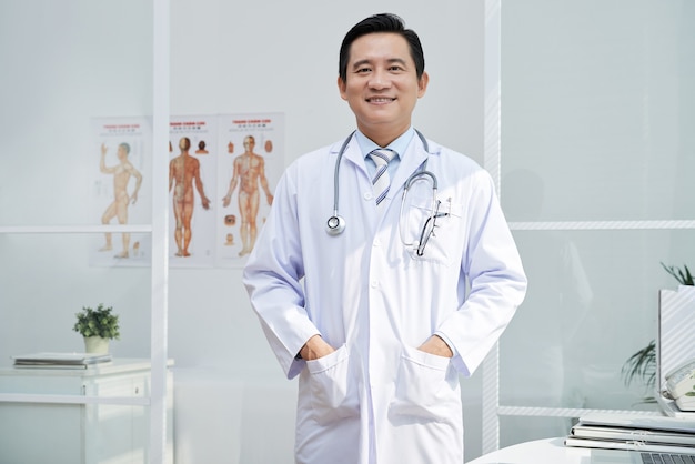 Portrait of mature general practitioner in labcoat standing with hands in pockets and smiling at camera