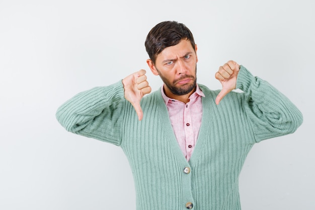 Portrait of mature man showing thumbs down in shirt, cardigan and looking grumpy front view