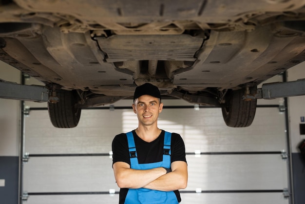 Portrait of mechanic repairing in uniform standing looking camera crossed hands at under lifted car