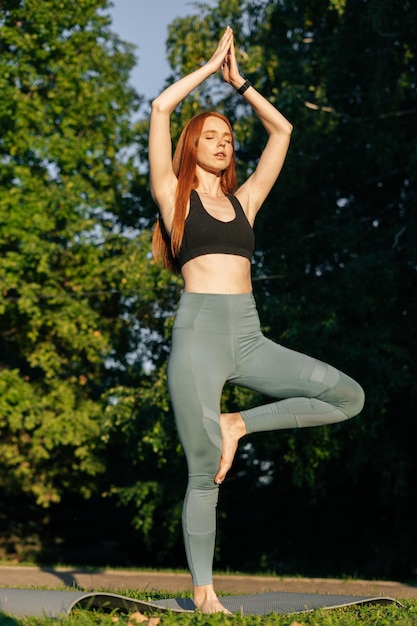 Portrait of meditative redhead young woman doing yoga in tree pose raising hands in Namaste pose