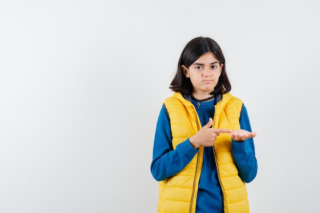 Portrait of mid cut hair teenager girl in blue sweater and yellow vest thinking and showing another hand isolated on white background