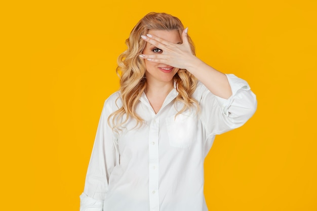 Portrait of a middle-aged European blonde woman in a white shirt, closes her eyes with her hands and looks through her fingers, smiling. Standing on a yellow background. A look into the future.