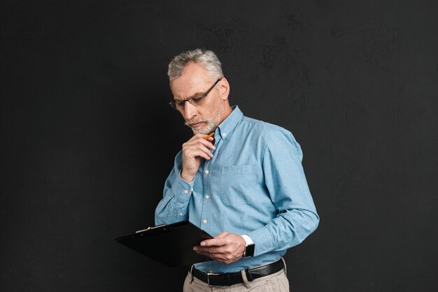 Portrait of middle aged man 60s with grey hair and beard working with documents while holding clipboard, isolated over black wall