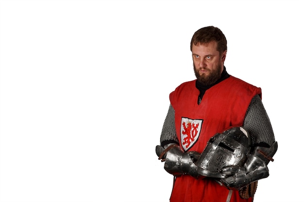 Portrait of a middle-aged man with a beard in the clothes and armor of a medieval knight with a helmet in his hands isolated on a white wall