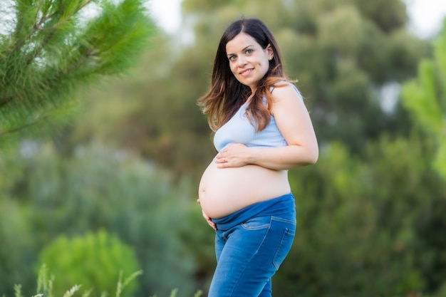 Portrait of a middle-aged pregnant latina woman in jeans standing in a park while touching her abdomen and looking at the camera.