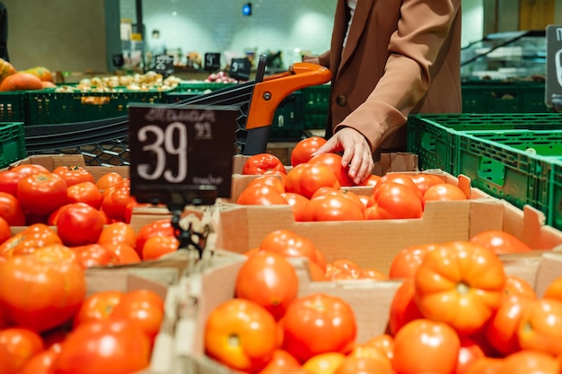 Portrait of a Millennial Lady buying food walking in a supermarket with a trolley A female customer chooses tomatoes taking products from the shelves