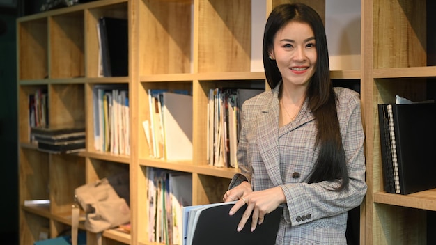 Portrait of millennial working woman in business suit standing near bookshelves in her personal office and smiling at camera