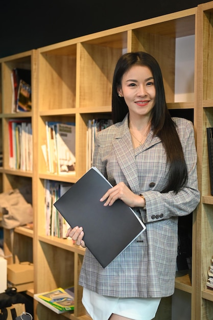 Portrait of millennial working woman standing near bookshelves in her personal office and smiling at camera