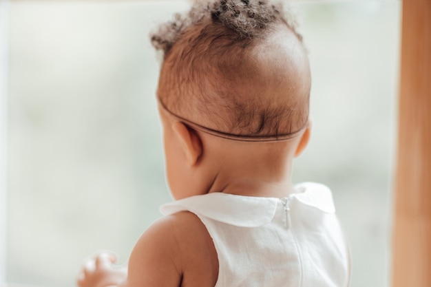 Portrait of mixed race toddler girl in white dress with baby toy sitting near window looking away