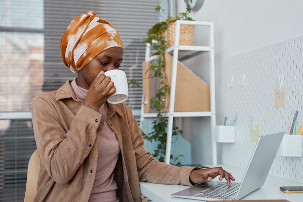 Portrait of modern African-American woman wearing headscarf in office and using laptop, copy space