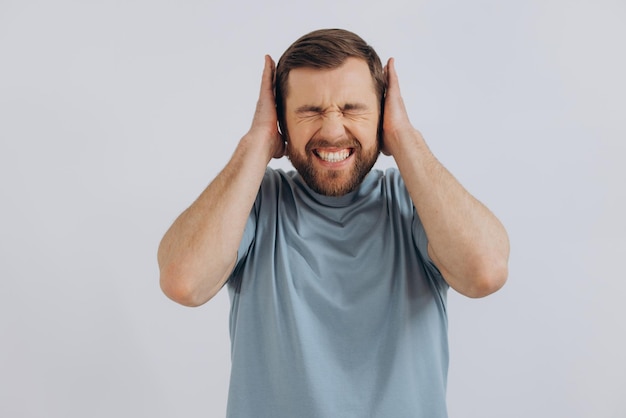 Portrait of a modern bearded middleaged man in a blue tshirt showing emotions covering his ears on a white background