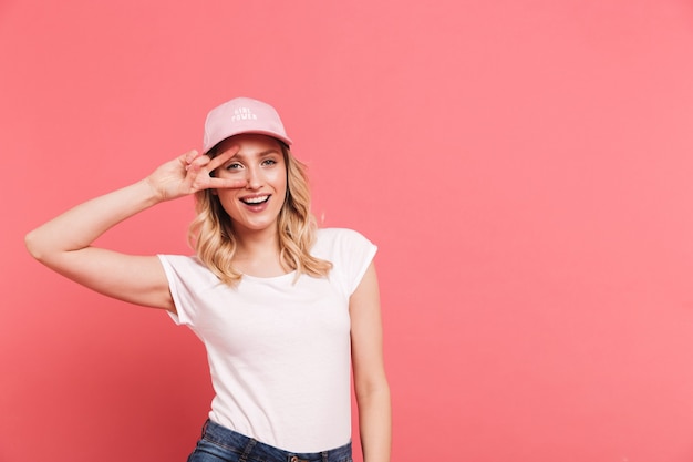 Portrait of modern curly woman wearing casual t-shirt and cap smiling and showing peace sign isolated over pink wall