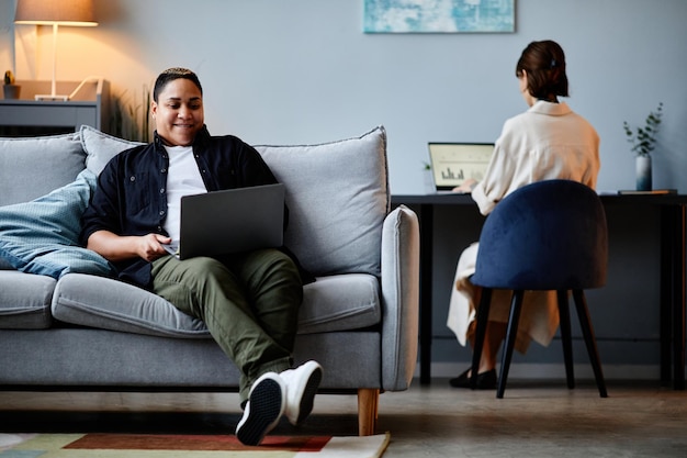 Portrait of modern multiethnic woman relaxing on couch at home and using laptop copy space