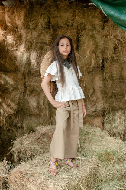 Portrait of modern stylish preteen brunette girl spending summer time in country estate posing on haystack in hayloft