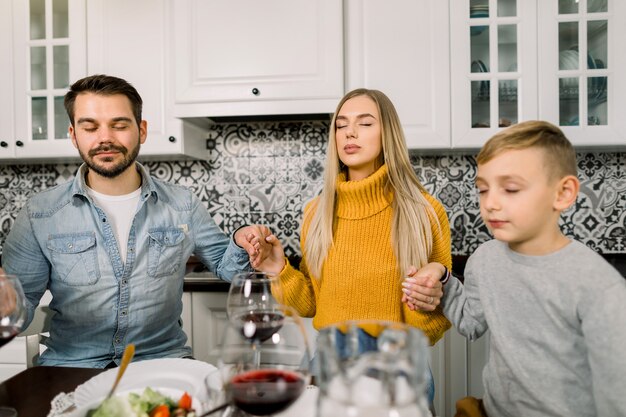 Portrait of modern young family, father, mother and son, sitting at festive table and praying.