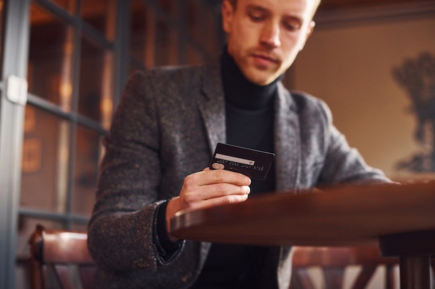 Portrait of modern young guy in formal clothes that sits in the cafe and holds credit card in hand.