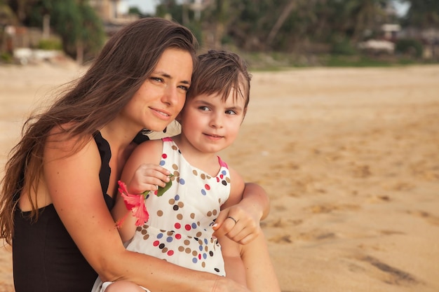 Portrait mother and little daughter together sitting on tropical sandy beach at palm trees background