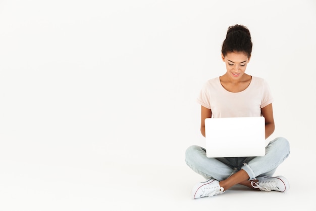 Portrait of mulatto woman in casual sitting on floor in lotus pose and holding laptop, isolated over white wall