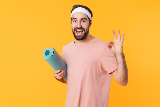 Portrait of muscular athletic young man in t-shirt smiling and carrying fitness mat isolated over yellow wall