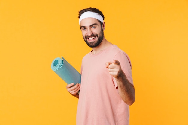 Portrait of muscular athletic young man in t-shirt smiling and carrying fitness mat isolated over yellow wall