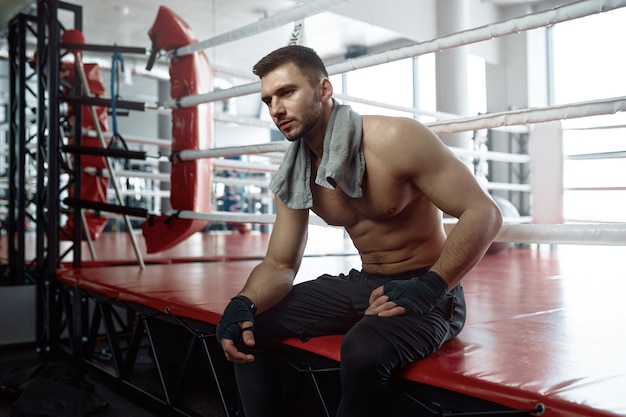 Portrait of muscular male boxer sitting on boxing ring mat and rest after hard training session or competitive match