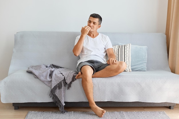 Portrait of nervous Caucasian handsome young adult man wearing white shirt and jeans short, biting his fingernails, posing indoor while sitting on gray sofa.