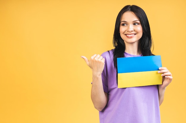 Portrait of nice beautiful lovely glad cheerful woman holding in hands Ukrainian flag having fun isolated over yellow pastel color background Pointing finger