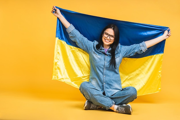 Portrait of nice beautiful lovely glad cheerful woman holding in hands Ukrainian flag having fun isolated over yellow pastel color background Sitting on the floor in lotus pose