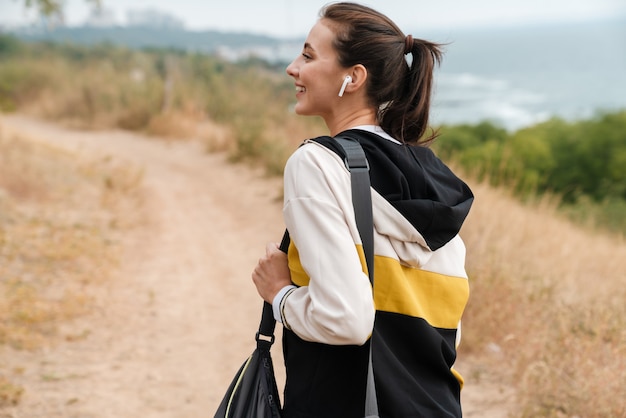 Portrait of nice laughing woman in sportswear using earpods while working with bag at seashore