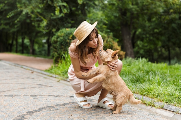 Portrait of nice woman wearing dress stroking dog and making kiss face while squatting in a park