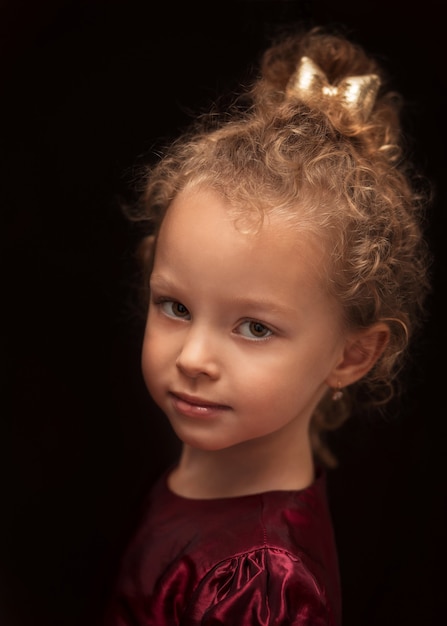 Portrait o beautiful little girl with curly hair and a bow in burgundy dress on a black background