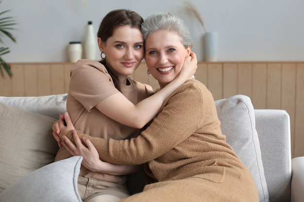 Portrait of old mother and mature daughter hugging at home. Happy senior mom and adult daughter embracing with love on sofa.