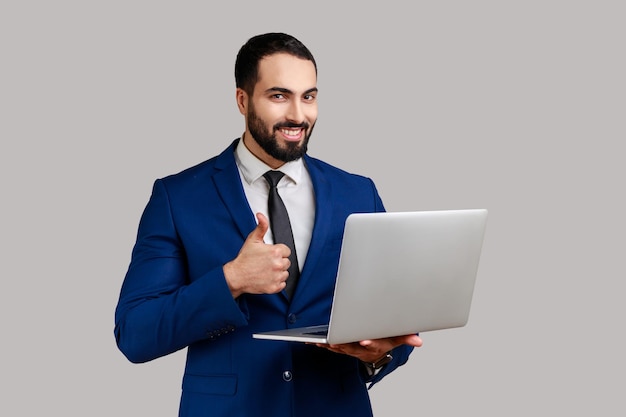 Portrait of optimistic bearded man holding laptop looking at camera and showing thumb up