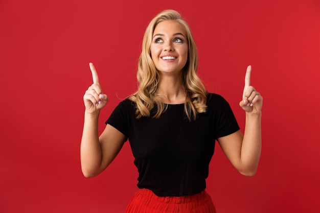 Portrait of optimistic blond woman 20s wearing red shirt pointing fingers upward on copyspace, isolated over red wall