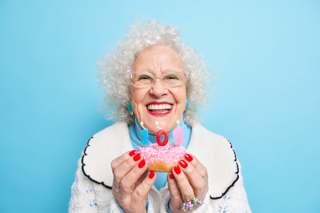 Portrait of optimistic curly haired woman holds delicious doughnut in hands smiles broadly has red nails enjoys birthday celebration blows number candles