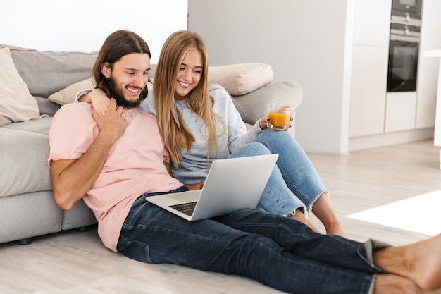 Portrait of optimistic hugging young loving couple near sofa using laptop computer indoors at home.