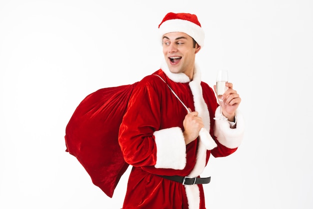 Portrait of optimistic man 30s in santa claus costume and red hat holding gift bag and glass with champagne