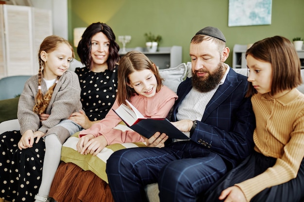 Portrait of orthodox jewish man wearing kippah while reading book to many children at home