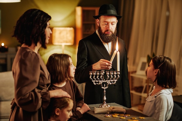 Portrait of orthodox jewish man with family lighting menorah candle during hanukkah celebration