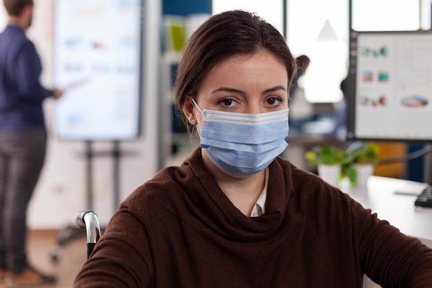 Portrait of paralyzed businesswoman wearing protective face mask to prevent infection with coronavirus working in startup business office. Manager in wheelchair analyzing financial charts